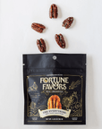 A black stand-up pouch of Fortune Favors 'The Everything' candied pecans packaging on a white background with scattered pecans.