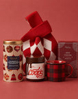 A holiday gift bag set featuring a Whittard cranberry and white chocolate chip biscuits, a jar of Coops peppermint hot fudge, a red and black buffalo plaid mug, a box of Gran Provisions hot cocoa and a red and white argyle knit bag, arranged on a solid red background.