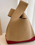 A knitted knot bag, featuring a khaki body and a contrasting red base, displayed on a white surface next to a book.