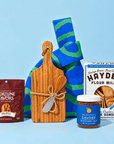 Gourmet gift collection featuring Fortune Favors spicy pecans, a bamboo cutting board with cheese knives, a blue and green patterned towel, Hayden Flour Mills crackers, and Momofuku savory seasoned salt, arranged against a light blue background.