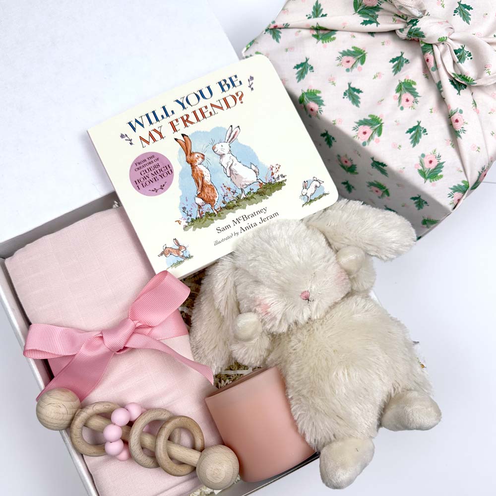 kadoo baby gift box with bunny stuffed animal, pink baby blanket, wooden rattle, pink candle, will you be my friend board book and more. Wrapped in rose pink fabric wrap.