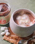 A ceramic mug filled with hot chocolate topped with marshmallows and a candy cane, next to an open jar of Coops's Peppermint Hot Fudge on a textured surface.