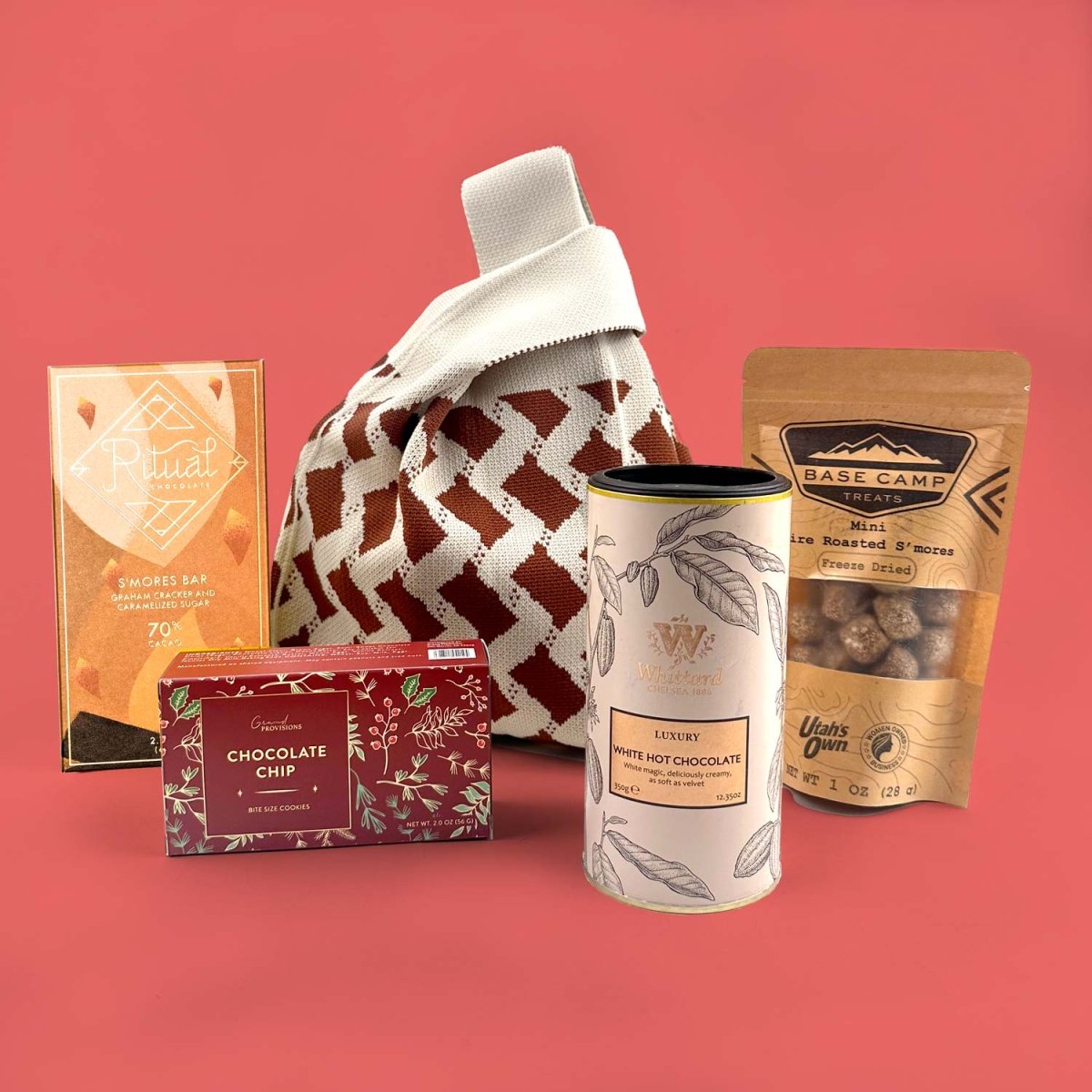 Collection of gift items including a hot chocolate mix, cookies, S'more chocolate bar, and roaster s'mores, knit bag,  on a red background.