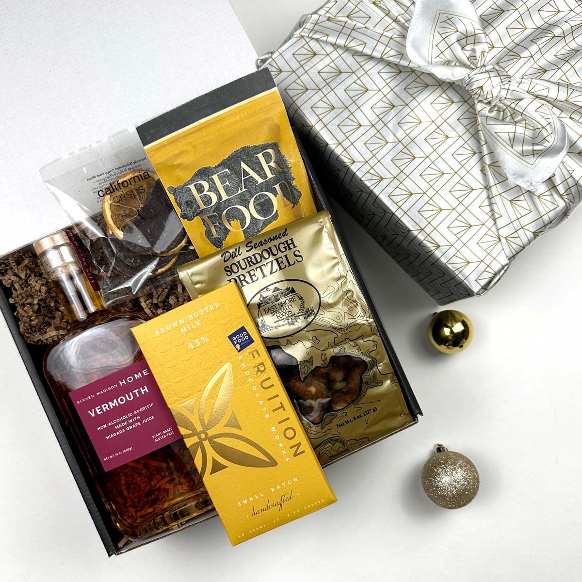 kadoo cocktail holiday gift box in white gold furoshiki wrap. Inside gifts: eleven madison home vermouth, fruition chocolate, bear food peanuts and more snacks