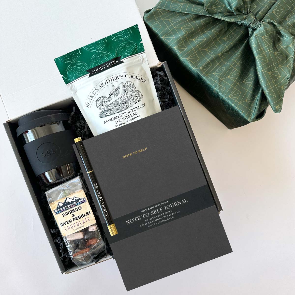 kadoo do great things gift box with black sol cup, shortbread, black notebook, pen, espresso beans chocolate and more. In Furoshiki fabric wrap.