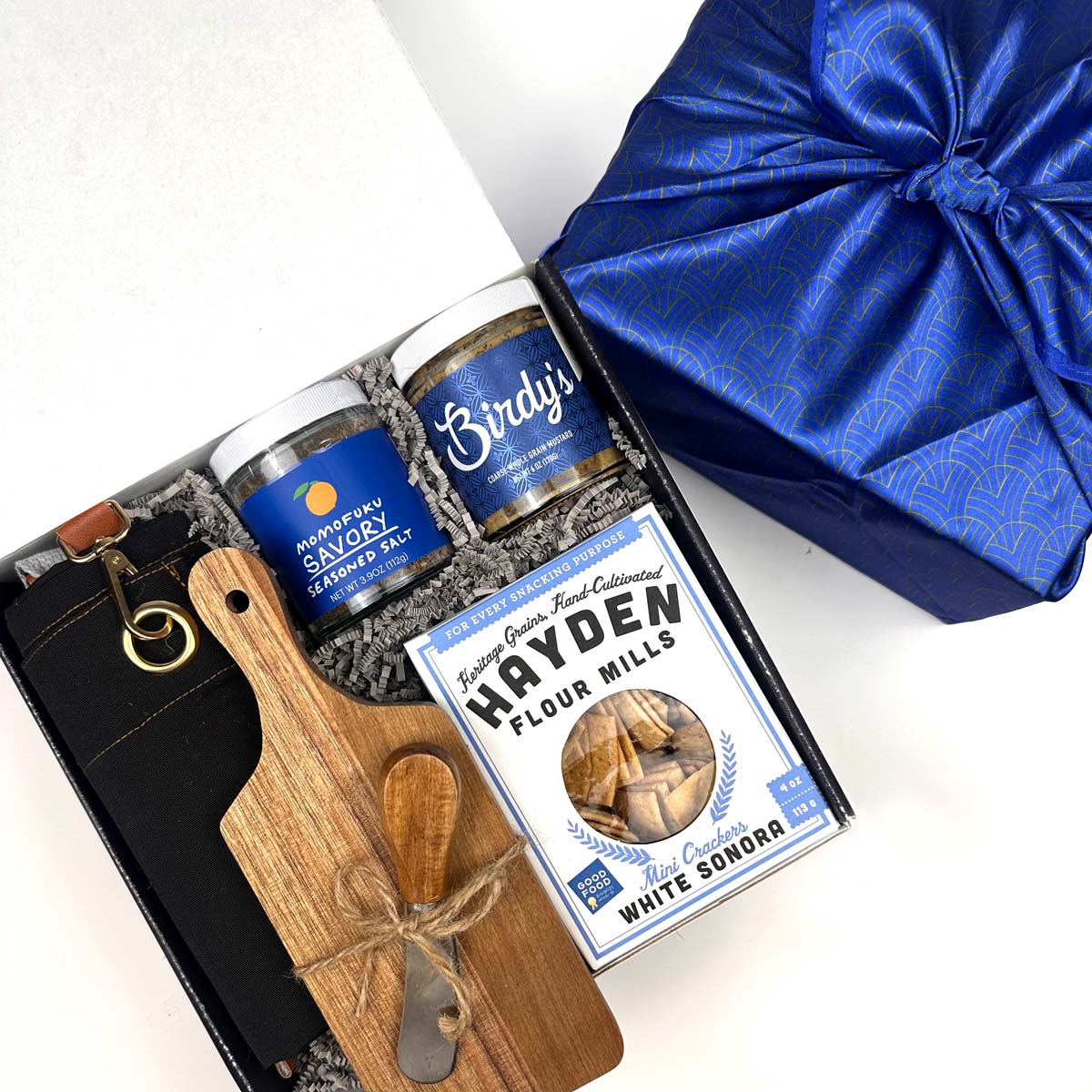 Gift set with momofuku's seasoned slat, Birdy's seasoning jars, a wooden cutting board, and a box of Hayden Flour Mills cracker, black apron, on a white background.