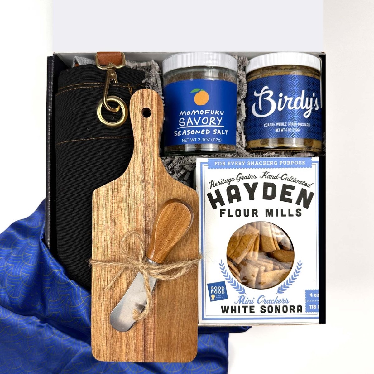 Set of gift items including a wooden cutting board with utensils, jars of seasoning salt, jar of mustard, a box of cracker, black folded apron on a blue fabric background.