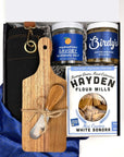 Set of gift items including a wooden cutting board with utensils, jars of seasoning salt, jar of mustard, a box of cracker, black folded apron on a blue fabric background.