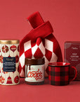 A holiday gift bag set featuring a Whittard cranberry and white chocolate chip biscuits, a jar of Coops peppermint hot fudge, a red and black buffalo plaid mug, a box of Gran Provisions hot cocoa and a red and white argyle knit bag, arranged on a solid red background.