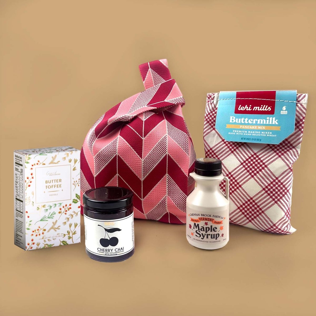 Collection of food and gift products including a jar of maple syrup, a bottle of jam, a box of chocolate, a bag of pancake and a pink bag, on a beige background