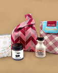 Collection of food and gift products including a jar of maple syrup, a bottle of jam, a box of chocolate, a bag of pancake and a pink bag, on a beige background