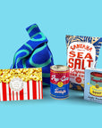Collection of snack items including a blue bag, popcorn box, truffle popcorn, sea salt chips, and Andy Warhol tomato soup can puzzle on a blue background.