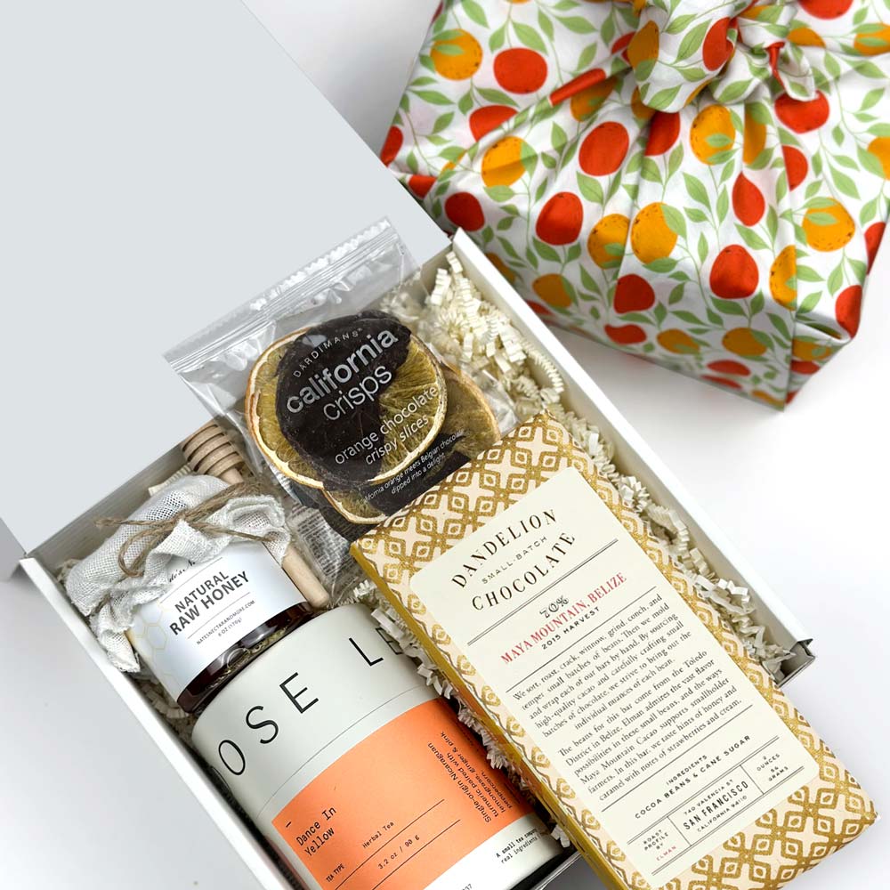 kadoo tea time curated gift wrapped in orange pattern fabric. Gift inside the box:  tea, natural raw honey, wooden honey dipper, dandelion chocolate bar, orange chocolate crisply slice and more.