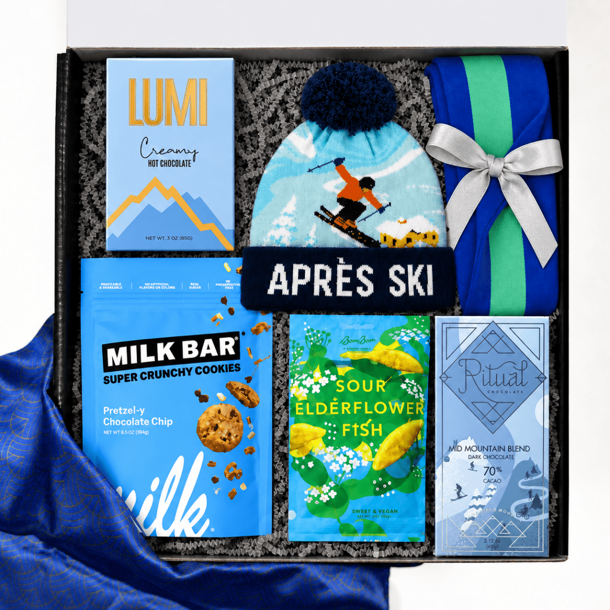 A curated gift box labeled “APRÈS SKI” containing a light blue ski-themed beanie with a dark pom-pom, a blue gift bag tied with a white ribbon, a MILK BAR chocolate bag, a LUMI hot chocolate box, a Ritual chocolate bar, and a colorful fish-themed card, all arranged in a black box.