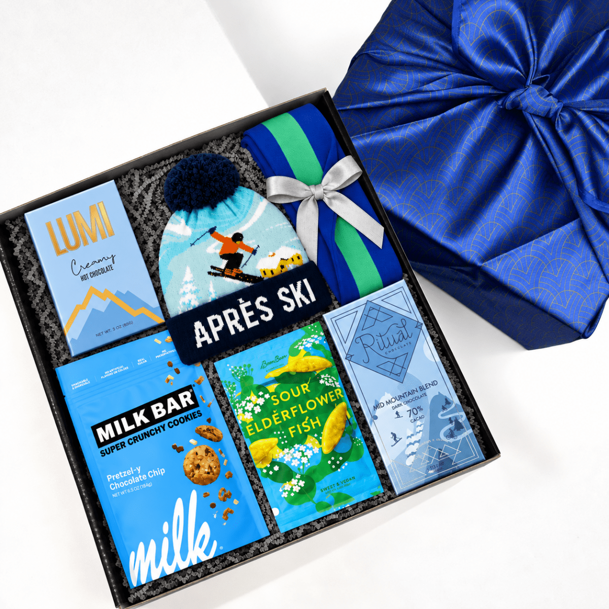 A curated gift box set labeled 'APRÈS SKI' arranged in a black tray, featuring a blue ski-themed beanie with a pom-pom, a striped blue and green towel, a Lumi snack box, a Milk Bar chocolate chip cookie box, a sour elderflower fish bar, and a Ritual peppermint candy box, next to a large blue satin gift box on a white background.