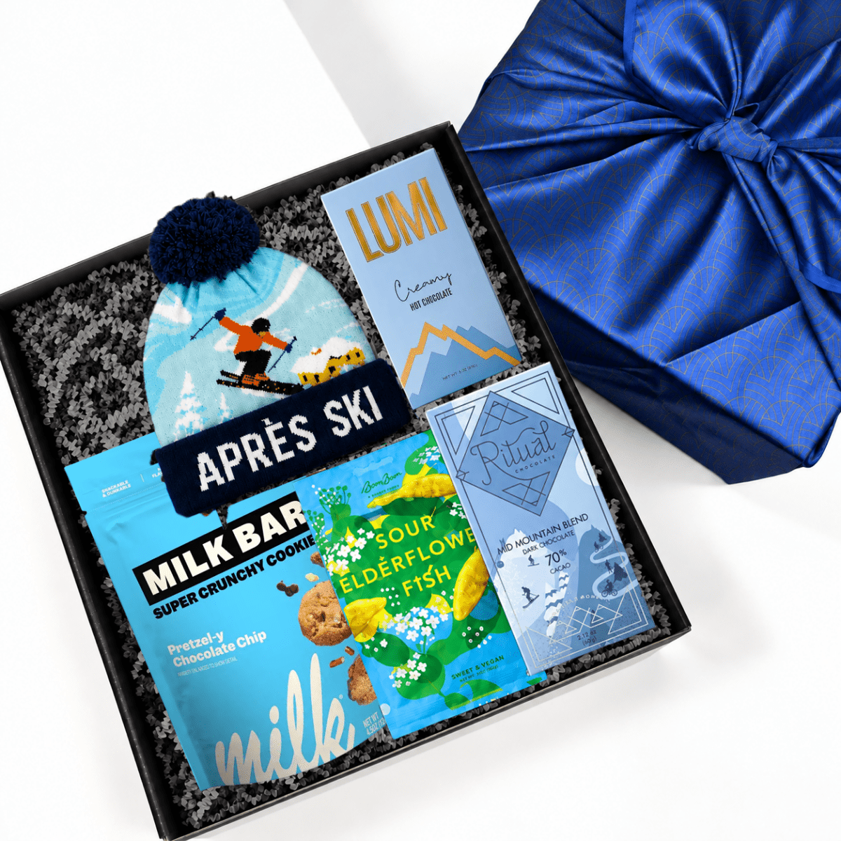 A curated gift box set containing an aprés ski snack bundle, including a milk bar, chocolate chip cookie, sour elderflower fish snack, and a blue pom-pom beanie, displayed next to a blue satin gift box.