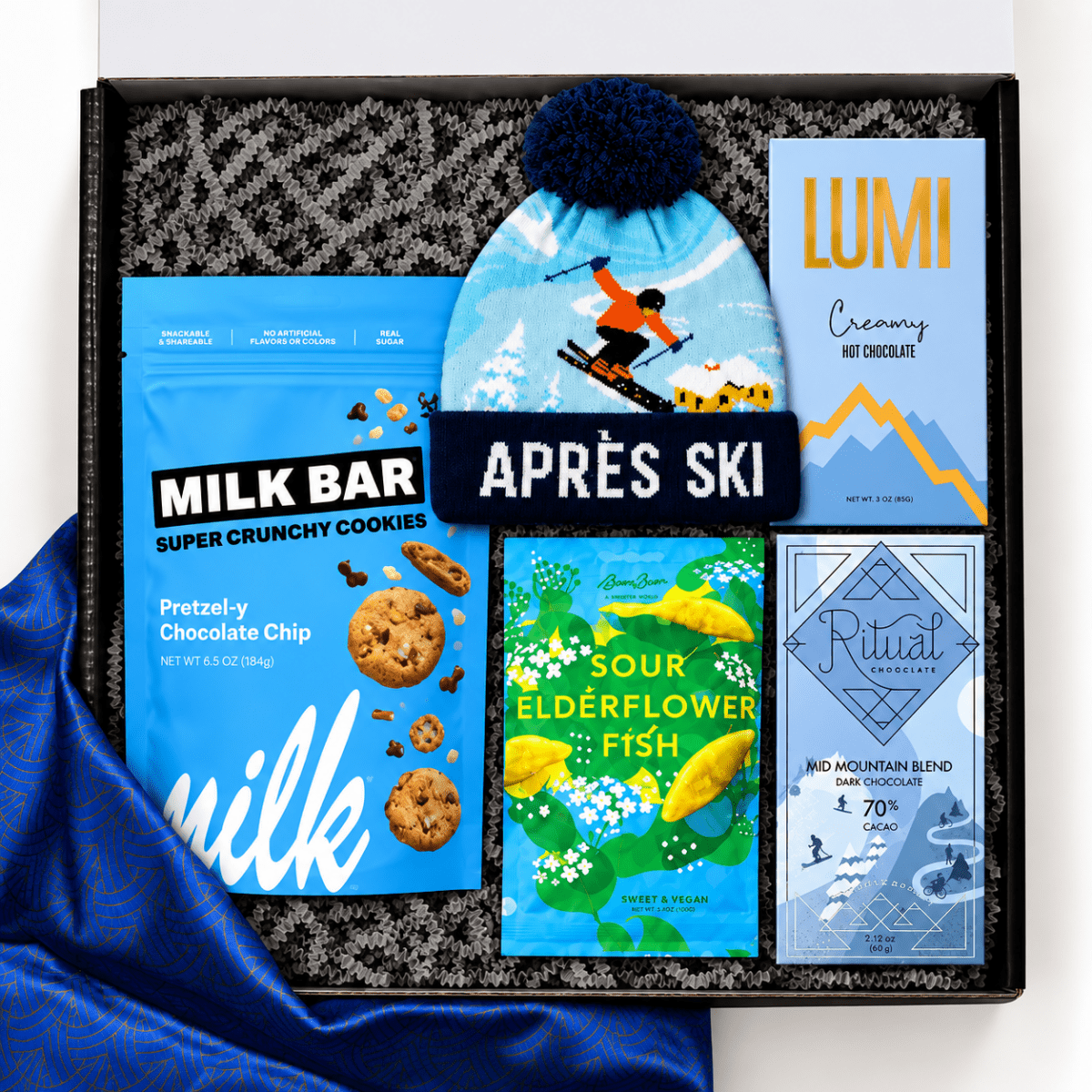 A curated gift box set containing a light blue beanie with a dark blue pom-pom and ski graphic, a blue bag of MILK BAR pretzel-y chocolate chip cookies, a blue and yellow APRÈS SKI hot chocolate box, a green and yellow Sour Elderflower Fish box, and a blue Ritual Mountains hot chocolate box, all arranged in a black gift box with gray patterned filler.