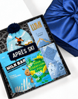 A curated gift box set containing an aprés ski snack bundle, including a milk bar, chocolate chip cookie, sour elderflower fish snack, and a blue pom-pom beanie, displayed next to a blue satin gift box.
