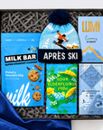 A curated gift box set containing a light blue beanie with a dark blue pom-pom and ski graphic, a blue bag of MILK BAR pretzel-y chocolate chip cookies, a blue and yellow APRÈS SKI hot chocolate box, a green and yellow Sour Elderflower Fish box, and a blue Ritual Mountains hot chocolate box, all arranged in a black gift box with gray patterned filler.
