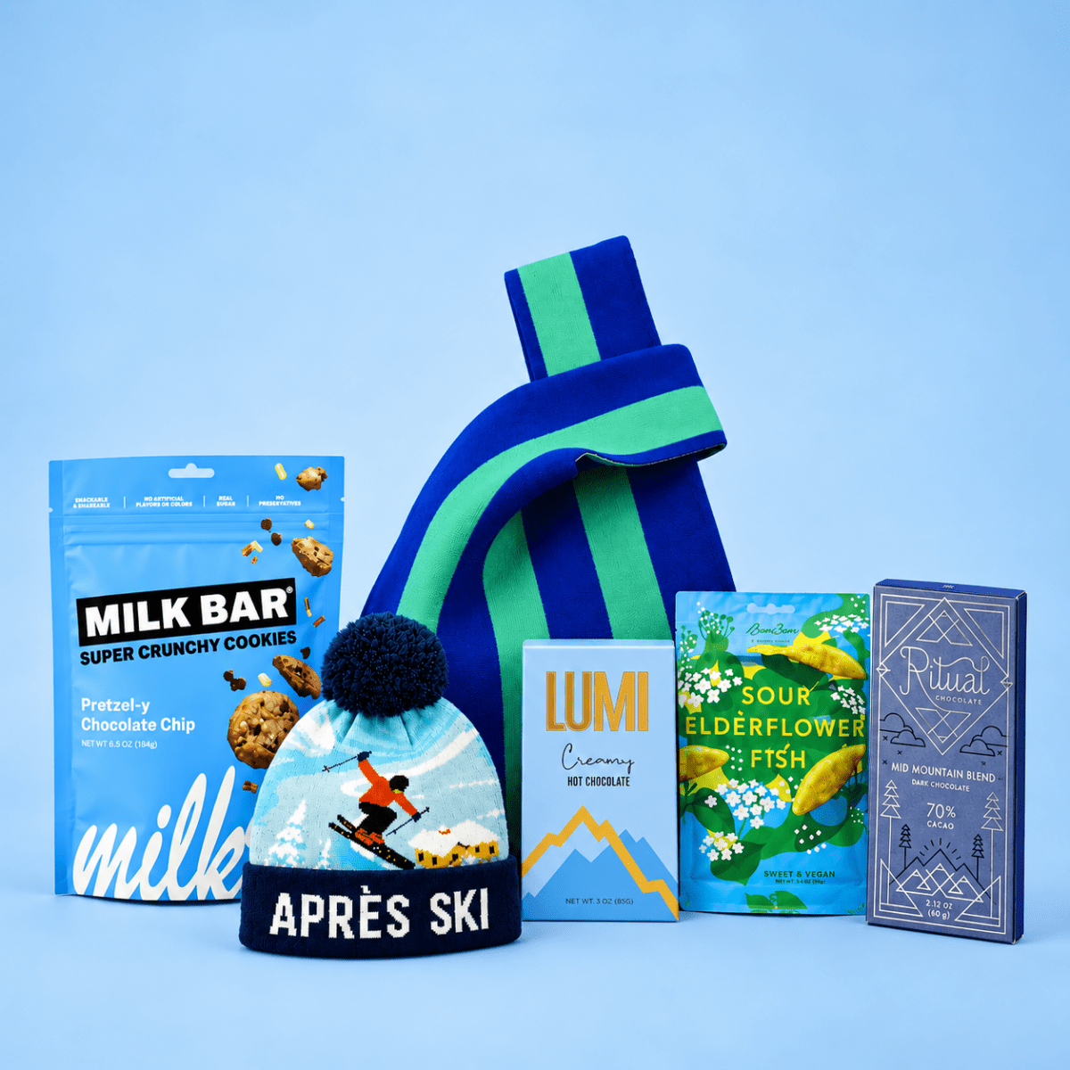 A curated gift set featuring a blue and green striped knit beanie, a bag of Milk Bar pretzels, a box of Lumi hot chocolate, a package of Sour Elderflower Fish, and a Ritual chocolate bar, arranged on a light blue background.