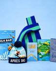 A curated gift set featuring a blue and green striped knit beanie, a bag of Milk Bar pretzels, a box of Lumi hot chocolate, a package of Sour Elderflower Fish, and a Ritual chocolate bar, arranged on a light blue background.