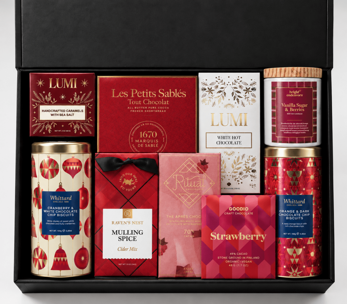 Open black holiday gift box filled with assorted gourmet chocolates, cookies, caramels, hot chocolate, and seasonal sweets in festive red, pink, gold, and cream packaging.