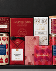 Open black holiday gift box filled with assorted gourmet chocolates, cookies, caramels, hot chocolate, and seasonal sweets in festive red, pink, gold, and cream packaging.