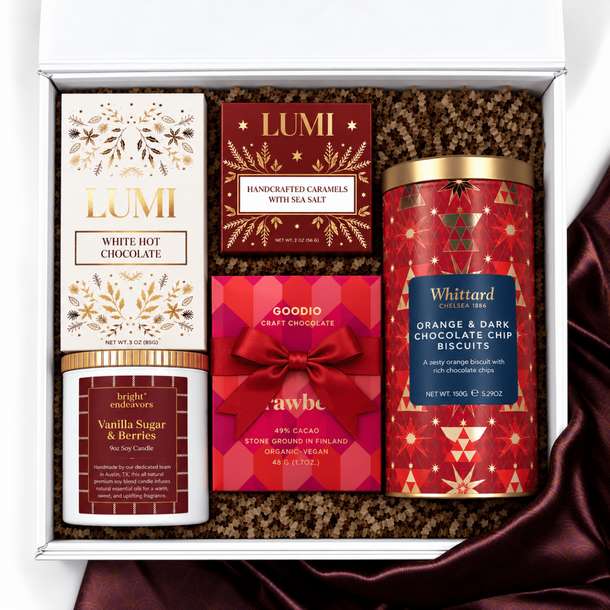 A curated gift box set containing multiple brands: Lumi White Hot Chocolate, Lumi Handcrafted Caramels, Goodio Craft Chocolate, and Whittard Biscuits, arranged in a white box with brown crinkle paper filler.
