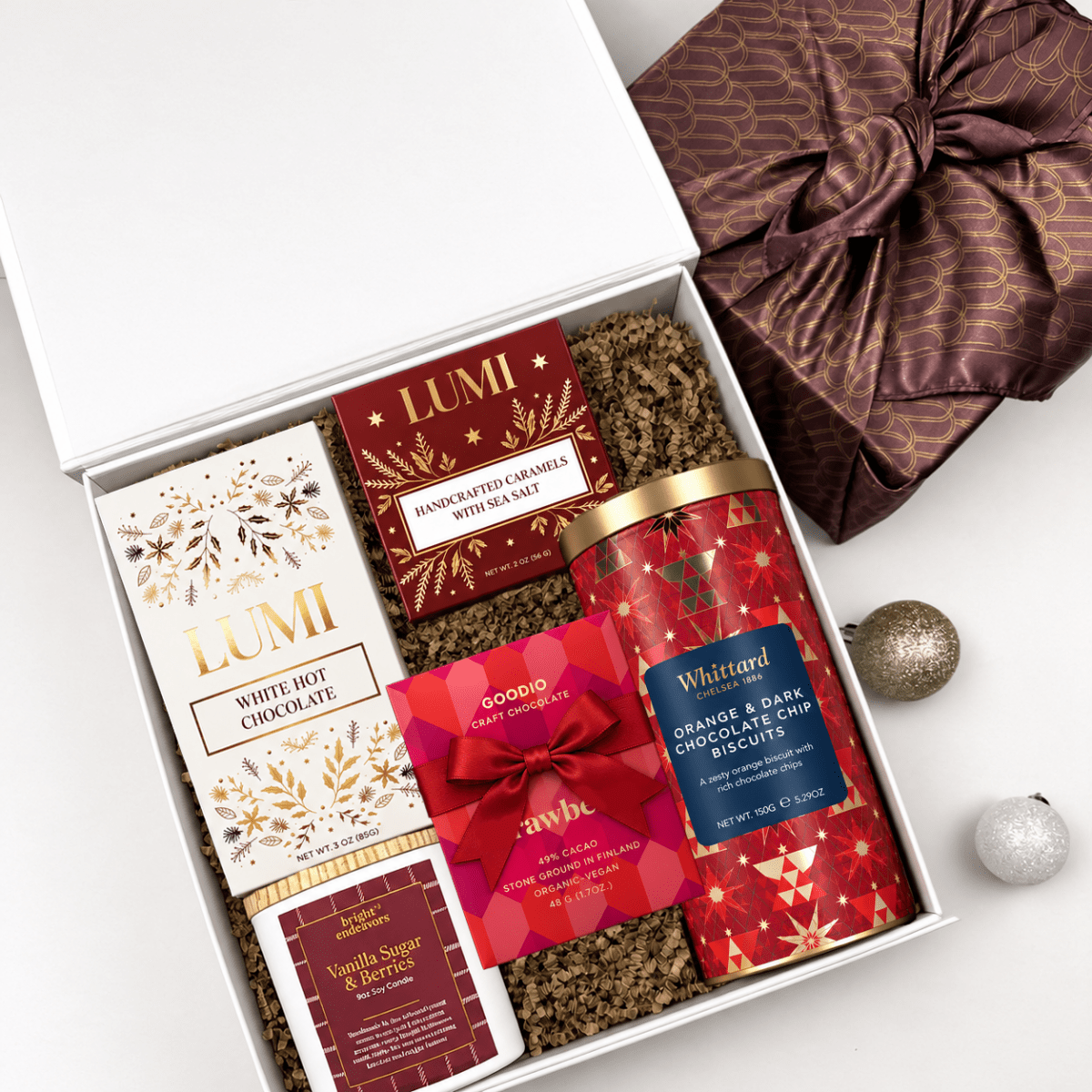An open white gift box filled with assorted gourmet chocolates and treats, styled on a light gray surface with a wrapped present, ornaments, and a bow. Visible products include Lumi White Hot Chocolate, Goodie's Vanilla Sugar & Barres, Wiltrand Orange & Dark Chocolate Candles, and another red gift-wrapped item.