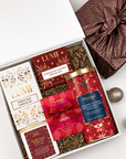 An open white gift box filled with assorted gourmet chocolates and treats, styled on a light gray surface with a wrapped present, ornaments, and a bow. Visible products include Lumi White Hot Chocolate, Goodie's Vanilla Sugar & Barres, Wiltrand Orange & Dark Chocolate Candles, and another red gift-wrapped item.