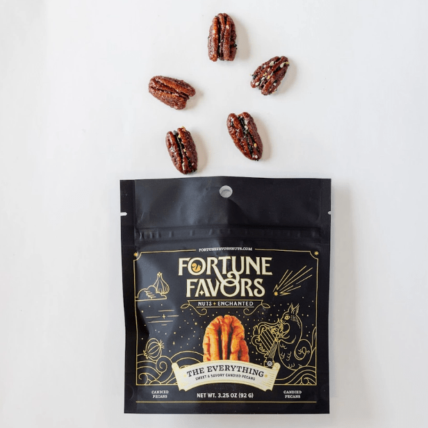 A black stand-up pouch of Fortune Favors 'The Everything' candied pecans packaging on a white background with scattered pecans.