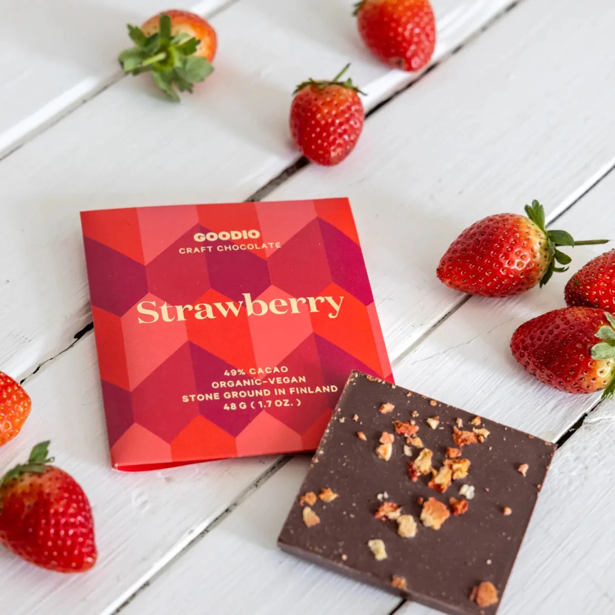 A package of Goodio Craft Chocolate Strawberry chocolate bar next to a square piece of dark chocolate topped with crushed nuts, styled on a white wooden surface with fresh strawberries.