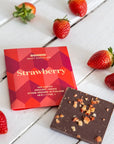 A package of Goodio Craft Chocolate Strawberry chocolate bar next to a square piece of dark chocolate topped with crushed nuts, styled on a white wooden surface with fresh strawberries.