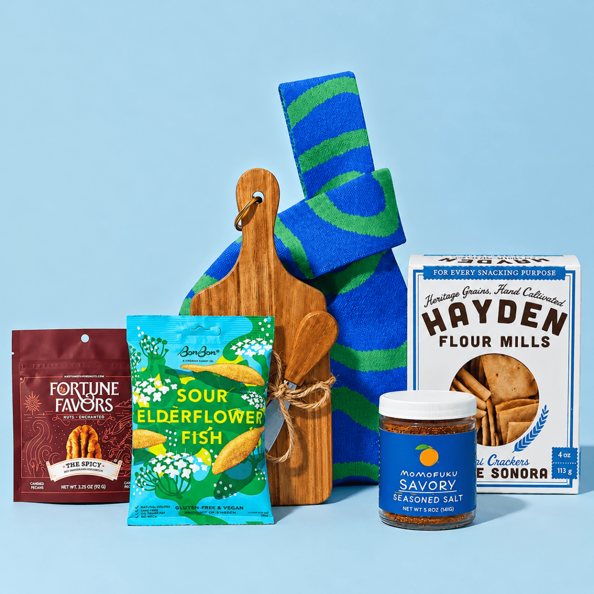 Gourmet gift assortment featuring Fortune Favors spicy pecans, BonBon Sour Elderflower Fish candy, Momofuku savory seasoned salt, Hayden Flour Mills crackers, a wooden cutting board with spreader, and a blue-and-green patterned towel, arranged against a light blue background