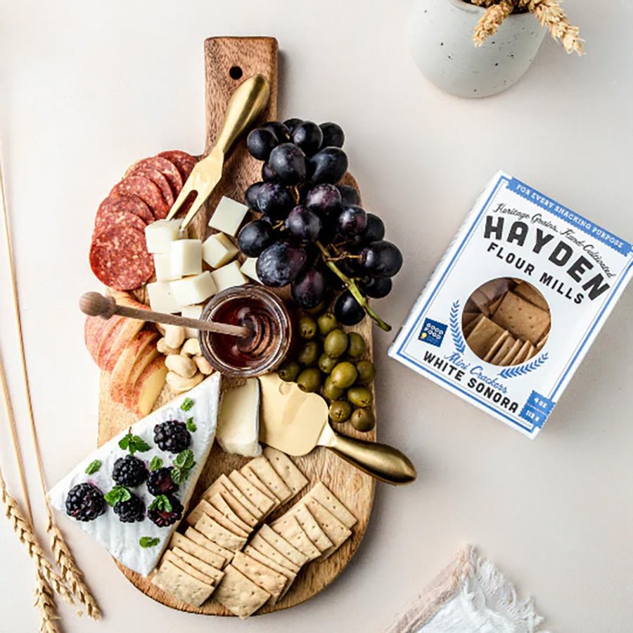 Wooden charcuterie board with assorted foods including cheese, fruits, and a box of Hayden Flour Mills white sonora crackers.