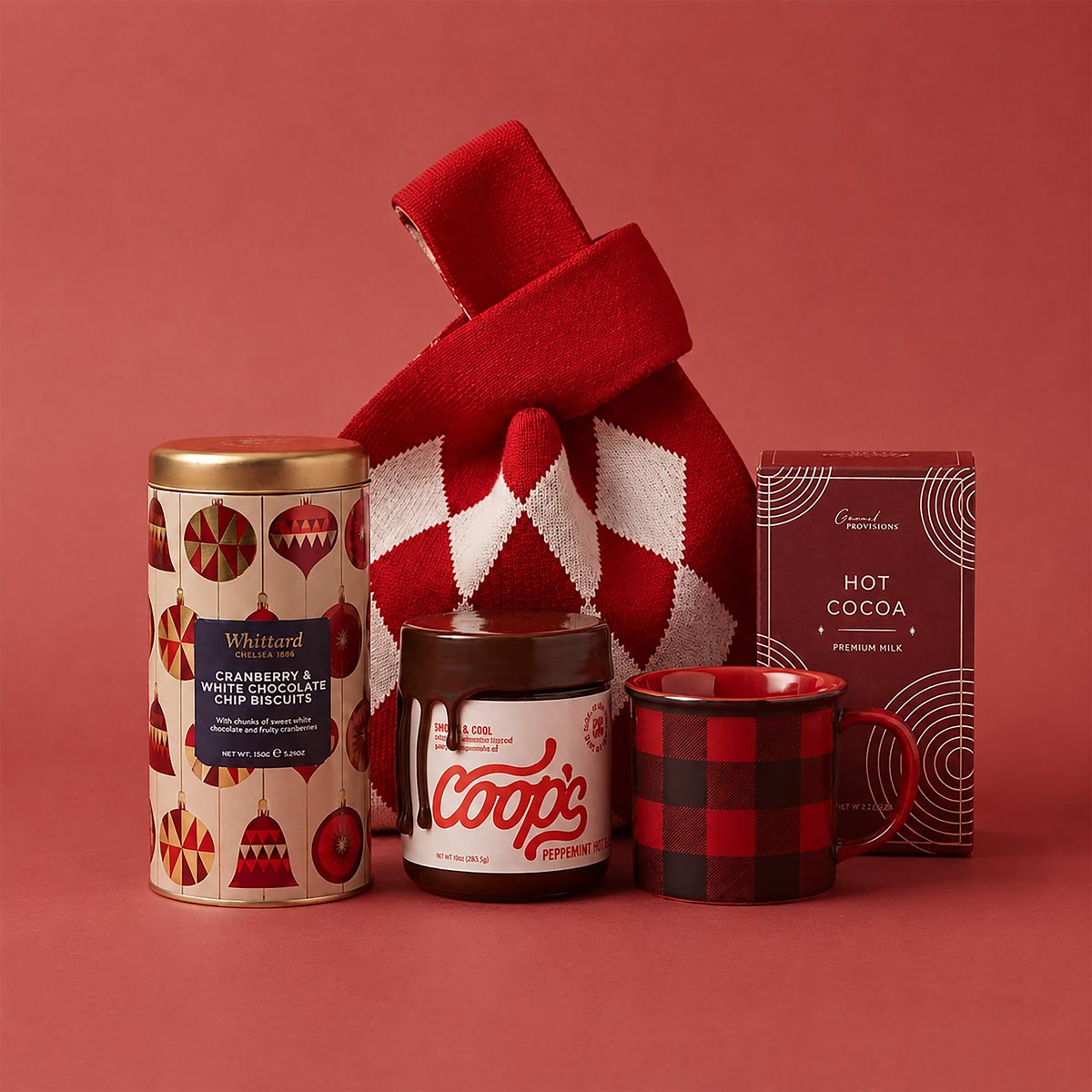 A holiday gift bag set featuring a Whittard cranberry and white chocolate chip biscuits, a jar of Coops peppermint hot fudge, a red and black buffalo plaid mug, a box of Gran Provisions hot cocoa and a red and white argyle knit bag, arranged on a solid red background.