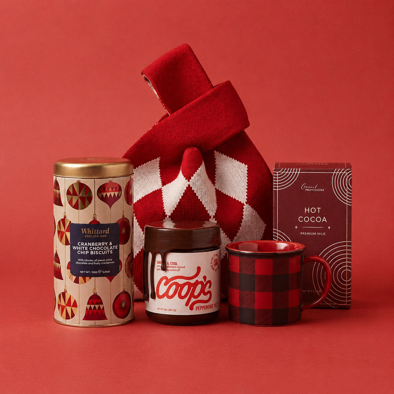 A holiday gift bag set featuring a tin of Whittard cranberry and white chocolate chip biscuits, a jar of Coops peppermint hot fudge, a red and black plaid stoneware mug, a red and white argyle knit bag, and a box of Grand Provision creamy milk hot chocolate arranged on a solid red background.