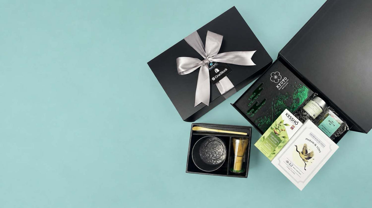 A curated wellness gift set arranged on a light aqua background, featuring a black presentation box with a silver bow, an open black box with green-patterned inserts, a small black tray with a gold item, and printed cards.