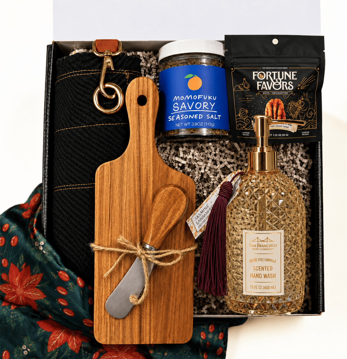 A curated gift box containing a wooden cutting board with a small knife tied in twine, a black knit sweater with a gold clasp, a jar of Savory Seasoned Salt, a bag of Fortune Favors tea, and a bottle of scented hand wash, arranged in patterned filler with a scarf underneath.
