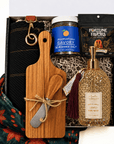 A curated gift box containing a wooden cutting board with a small knife tied in twine, a black knit sweater with a gold clasp, a jar of Savory Seasoned Salt, a bag of Fortune Favors tea, and a bottle of scented hand wash, arranged in patterned filler with a scarf underneath.
