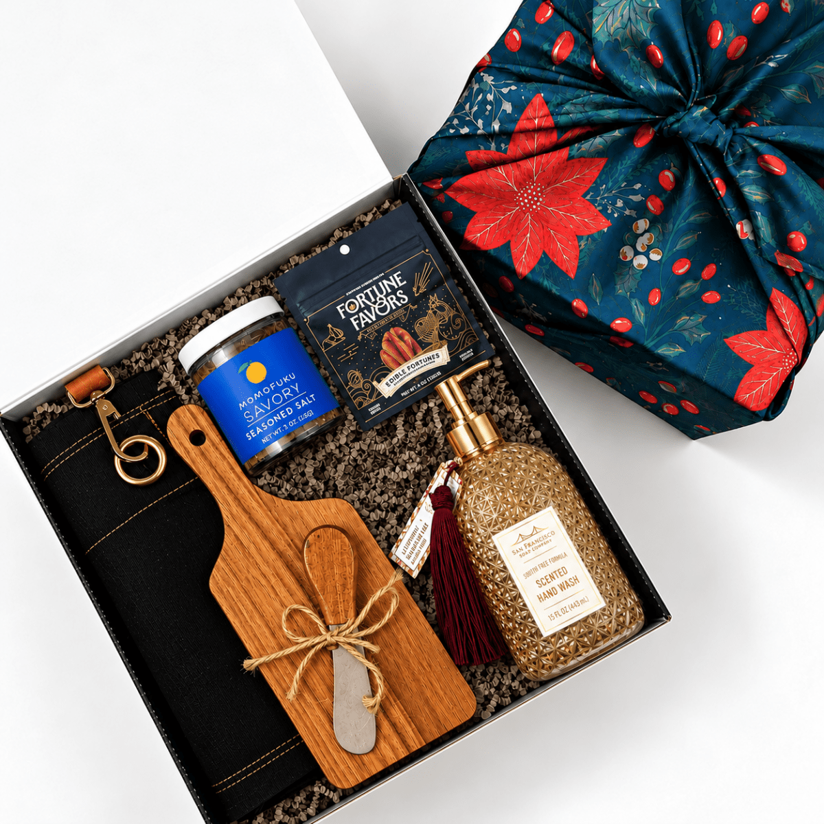 A curated gift box set shown from above on a white background. The open black gift box contains a wooden cutting board with a wooden spoon, a jar of seasoning, a black pouch of Fortune Favors, and a clear bottle of hand wash.