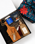 A curated gift box set shown from above on a white background. The open black gift box contains a wooden cutting board with a wooden spoon, a jar of seasoning, a black pouch of Fortune Favors, and a clear bottle of hand wash.