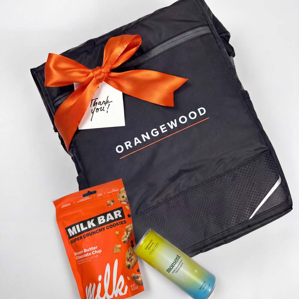 Black Orangewood bag with an orange ribbon and gift tag, accompanied by a milk chocolate bar and a can of mountain dew.