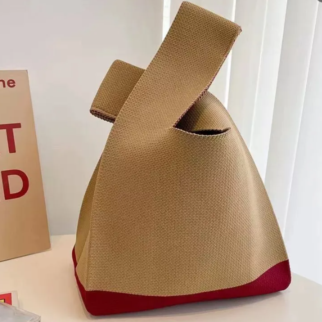 A knitted knot bag, featuring a khaki body and a contrasting red base, displayed on a white surface next to a book.