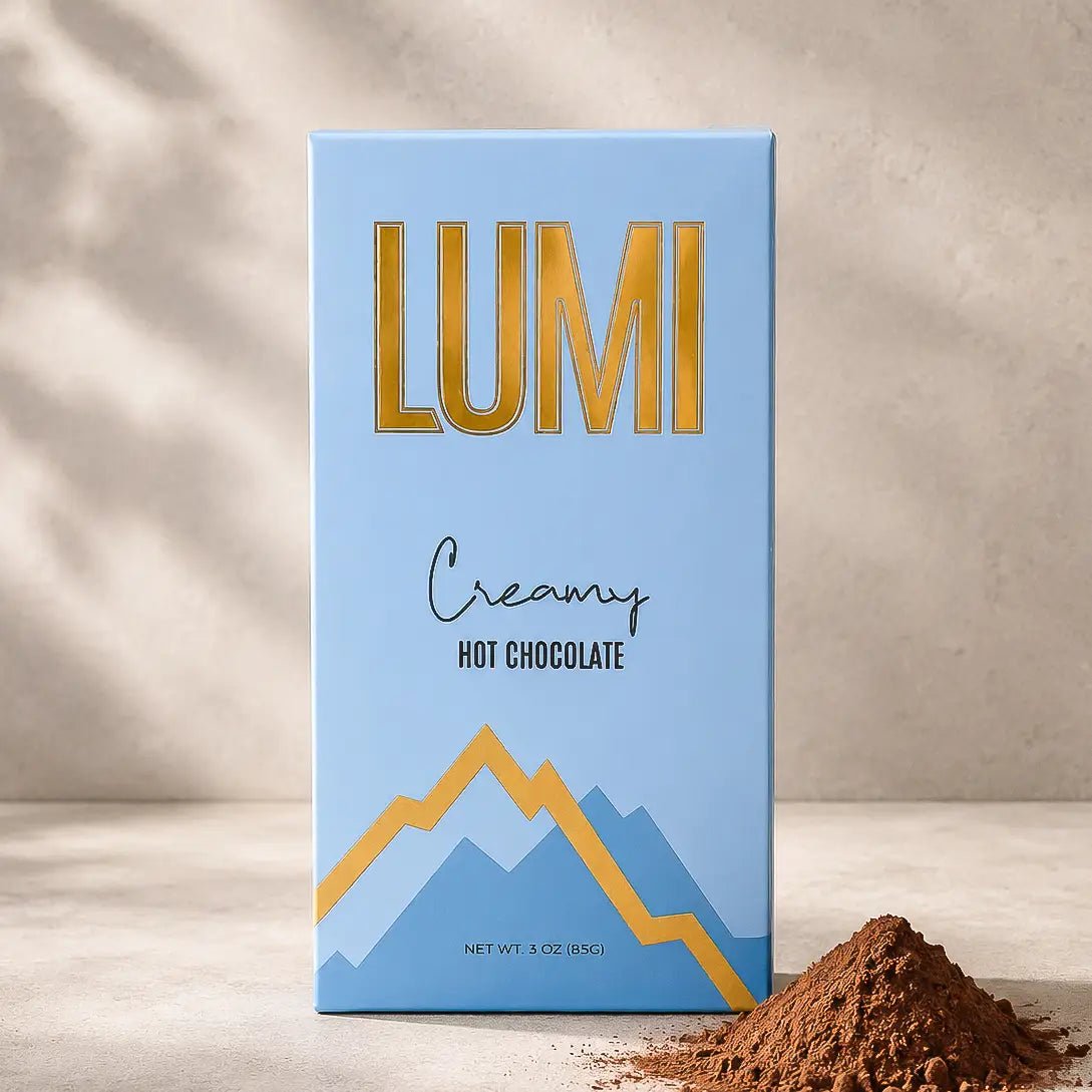 A rectangular box of Lumi Creamy Hot Chocolate stands upright on a light surface against a textured beige wall with soft shadow overlays. The box is light blue with large gold 'LUMI' lettering, smaller black script text, and a blue-and-gold mountain graphic.