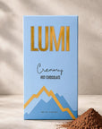 A rectangular box of Lumi Creamy Hot Chocolate stands upright on a light surface against a textured beige wall with soft shadow overlays. The box is light blue with large gold 'LUMI' lettering, smaller black script text, and a blue-and-gold mountain graphic.
