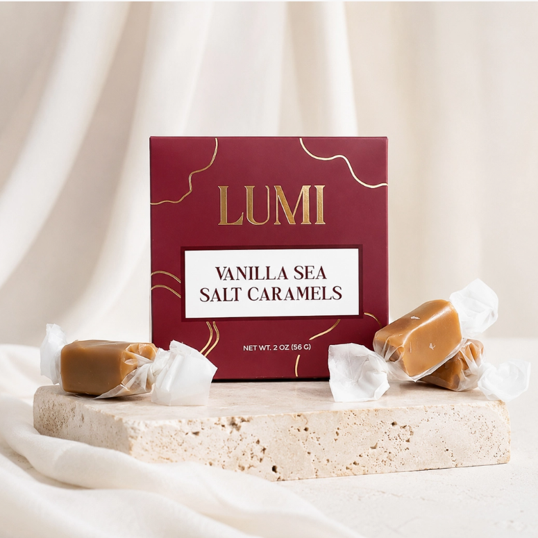 A burgundy color box of LUMI Vanilla Sea Salt Caramels stands on a stone slab with several wrapped caramels placed around it against a soft cream fabric backdrop.
