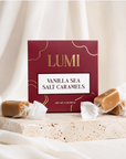 A burgundy color box of LUMI Vanilla Sea Salt Caramels stands on a stone slab with several wrapped caramels placed around it against a soft cream fabric backdrop.