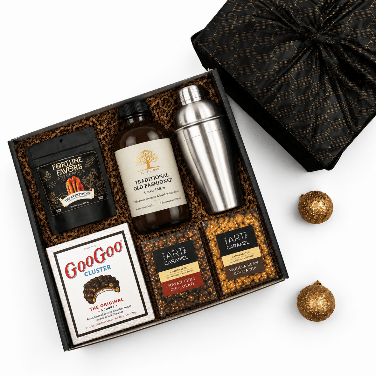A curated gift box set on a white background, featuring a black and gold patterned gift box with its lid off, containing a bottle of Rustic Oak Traditional Old Fashioned Mix, a stainless steel shaker, a bag of Fortune Favors Peanuts, a cookie box, and two bags of popocorns. Two gold decorative ornaments are placed beside the box.