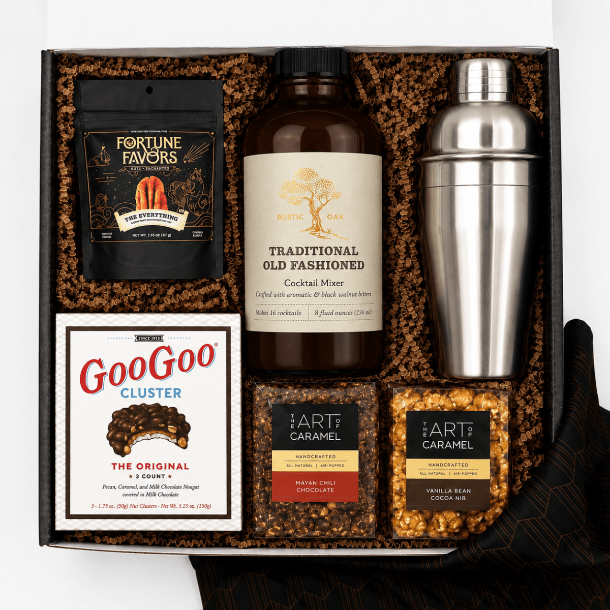 A curated gift box set containing a white bottle of Rustic Oak Old Fashioned Cocktail Mixer, a stainless steel shaker, a bag of Fortune Favors peanuts, a box of GooGoo Cluster chocolate, and two bags of Art of Caramel snacks, arranged in brown crinkle paper inside a black box.